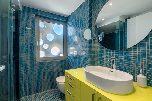 Koupelny jsou výrazně barevné – modré obklady kontrastují se zářivě žlutými skříňkami. Za oknem je viditelná venkovní stěna s opakujícím se motivem kruhových výřezů. 