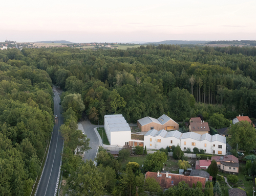 Pozemek pro výstavbu souboru budov je z jedné strany krytý lesem, z druhé navazuje na drobnou zástavbu.